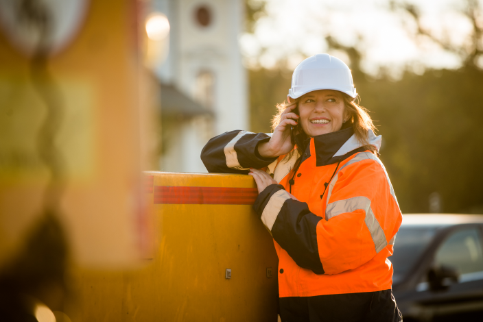 Female technician wearing high vis and hard hat talking on mobile phone, leaning on a front end loader