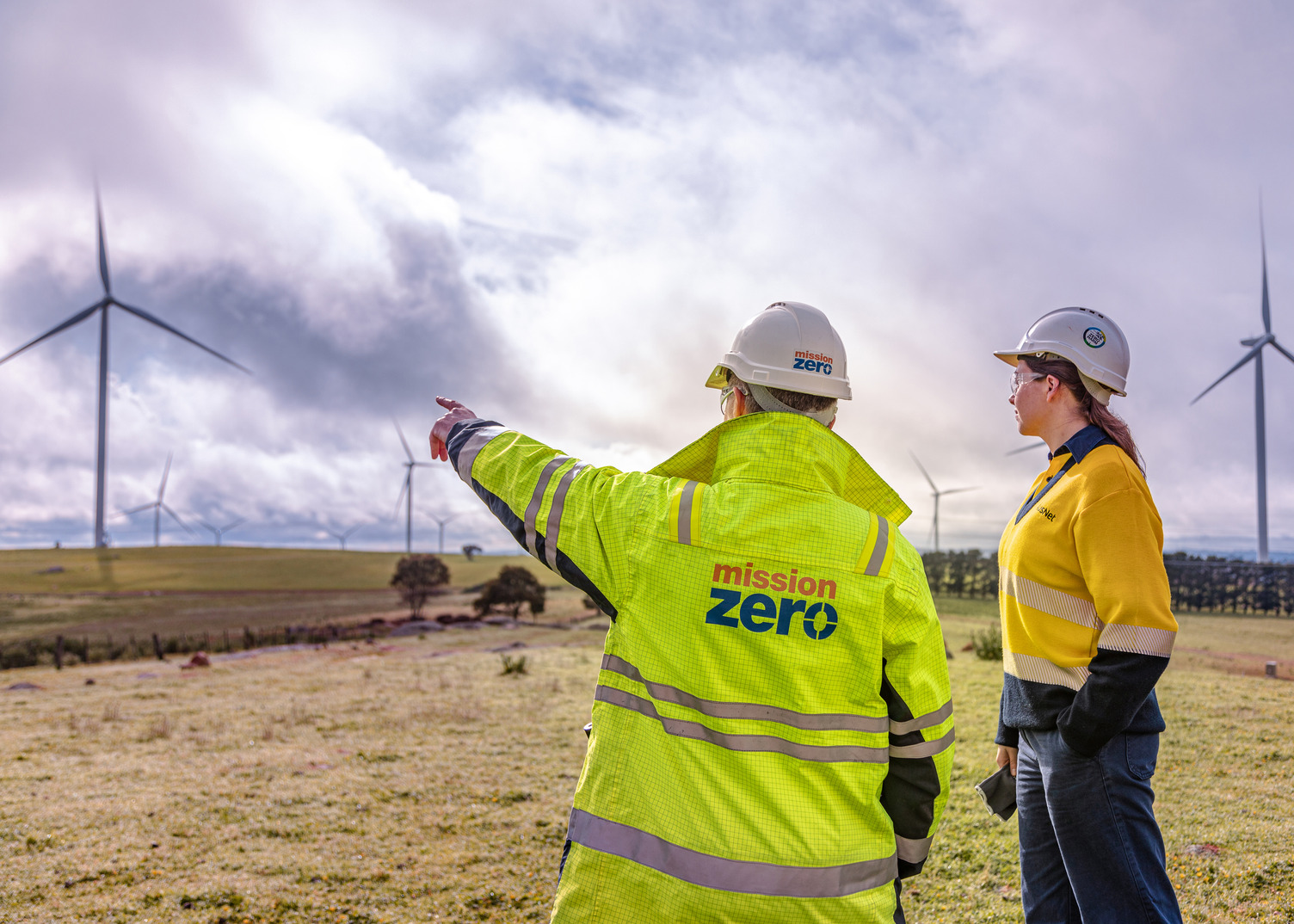 Two workers in high vis uniform pointing to wind farms in an open field