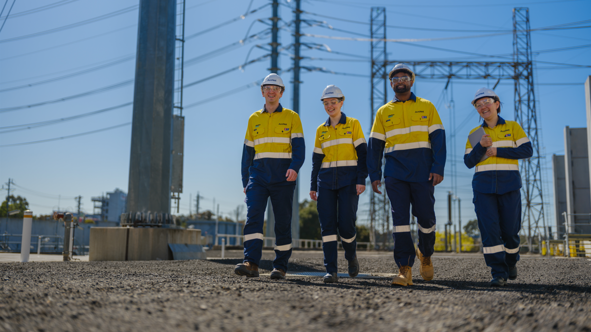 Four people walking outside in high-vis near an electricity substation