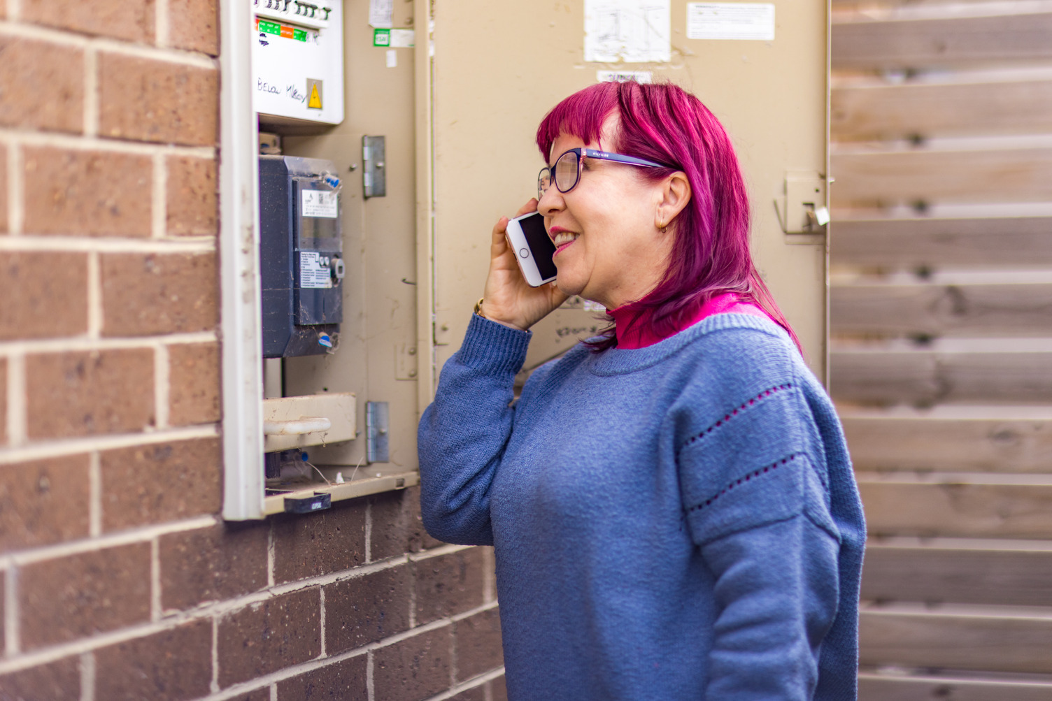 A woman with pink hair talking on a phone near an electric fuse box
