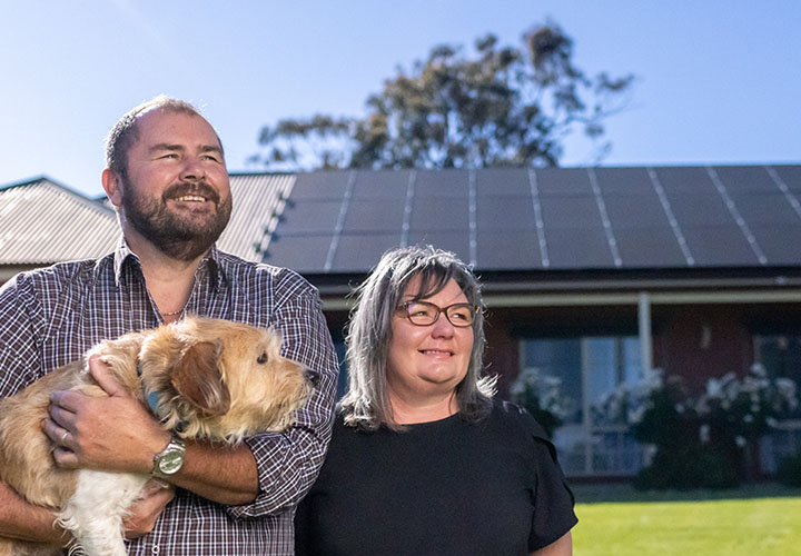 A smiling man holding a dog and smiling woman outside infront of solar panels on a roof