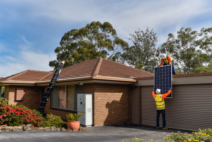 Single story house on a sunny day with two technicians wearing high vis and hard hats installing solar panels 